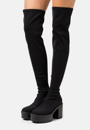 Schwarze Overknee-Plattformstiefel mit klobigen hohen Absätzen und seitlichen Reißverschlüssen, getragen an nackten Beinen vor einem schlichten Hintergrund.