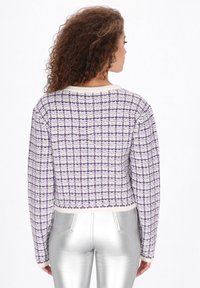 Rövidített kötött pulóver fehér és lila kockás mintával, hosszú ujjal és kerek nyakkivágással. Fényes ezüst nadrággal párosítva.