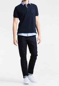 Camisa de polo navy con el logo H, cuello azul claro, combinada con jeans de mezclilla oscuros y zapatillas blancas, de pie contra un fondo liso.