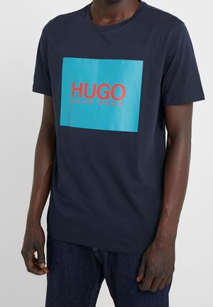 T-shirt bleu marine à manches courtes présentant un grand graphique rectangulaire avec "HUGO" en rouge sur un fond turquoise, fabriqué en coton.