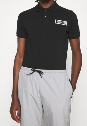 Personne portant un polo noir à manches courtes avec le logo « ICEBERG » et un pantalon gris clair à cordon de serrage avec des poches zippées.