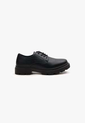 Zapato negro de cuero con cordones, punta redonda, suela gruesa y con taqueado, y textura suave; presenta costuras mínimas y un diseño sencillo.