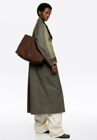 Ilgas alyvuogių spalvos paltas su rudo atspalvio akcentais, dėvimas ant kreminių kelnių ir juodų batų. Ruda rankinė su logotipu, modelis stovi šonu.