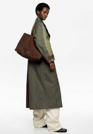 Długi oliwkowozielony płaszcz z brązowymi akcentami, noszony na kremowych spodniach i czarnych butach. Brązowa torba na ramię z logo, model stojący z boku.