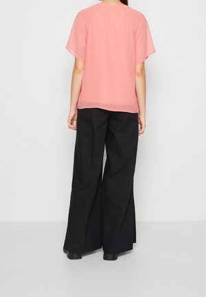 Persona de pie de espaldas, con una blusa rosa holgada de manga corta y pantalones anchos negros sobre un fondo claro y liso.