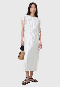 Abito midi bianco con maniche corte in chiffon, cintura regolabile con cordino e dettagli in pizzo. Modella che tiene una borsa di paglia, indossando sandali neri.