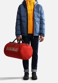 Puffy blue jacket, yellow fleece hoodie, dark jeans, black shoes; holding a red duffel bag with "NAPAPIJRI" logo and brown handles.