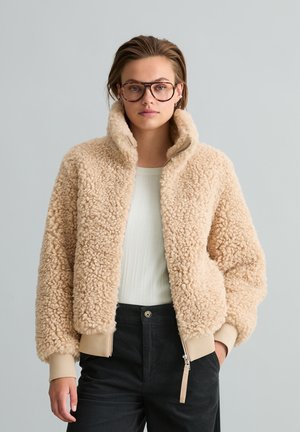 Donna con grandi occhiali tartarugati e una giacca beige soffice con zip, con pantaloni neri su sfondo grigio.