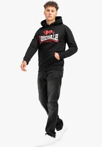 Sudadera negra con logo "Lonsdale" en rojo y blanco, bolsillo tipo canguro, capucha con cordón, combinada con jeans negros y zapatillas blancas.
