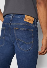 Vue de dos d'une personne portant un jean bleu Lee avec un écusson en cuir beige et des surpiqûres jaunes sur les poches, associé à une chemise gris foncé.