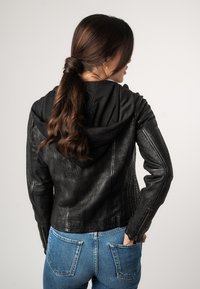 Schwarze Lederjacke mit gerippter Textur und Reißverschlussärmeln, ausgestattet mit einer schwarzen Kapuze. Getragen über blauen Denim-Jeans. Rückansicht.