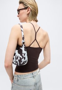 Γυναίκα με κοντά ξανθά μαλλιά, φοράει γυαλιά ηλίου, καφέ τοπ με λουριά, ανοιχτόχρωμο τζιν και κρατάει μαύρη και λευκή τσάντα ώμου με σχέδιο.