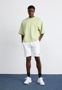 Lys grøn oversized t-shirt, hvide shorts med snørrebånd og hvide sneakers med teksturerede såler. Simpelt design, afslappet pasform.