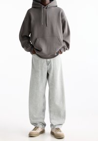 Person wearing oversized gray hoodie, loose light-wash jeans, beige sneakers, standing with hands in hoodie pocket against white background.