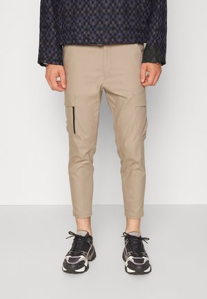 Beige cargobroeken met taps toelopende benen, twee zijzakken en een ritszak aan de linker dij. Draag ze met zwart-witte sportschoenen.