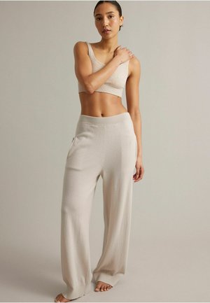 Femme debout pieds nus portant un crop top beige sans manches et un pantalon large en maille assorti sur fond uni.