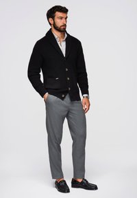 Czarny dzianinowy kardigan z kołnierzem szalowym, z drewnianymi guzikami i dwiema przednimi kieszeniami, zestawiony z szarymi dopasowanymi spodniami i czarnymi butami.