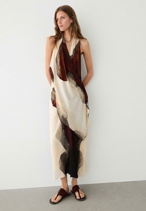 Femme portant une robe maxi sans manches crème et marron à motif abstrait avec un décolleté plongeant en V et des sandales marron, debout devant un mur blanc uni.