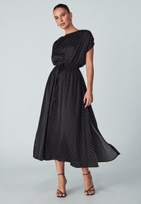 Black polka dot maxi dress with elastic waist, short sleeves, and flowing fabric. Accompanied by black strappy heels.