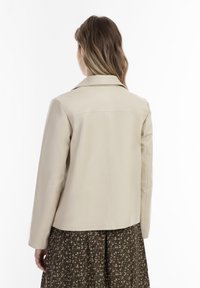 Beige leren jas met een kraag, een getailleerde pasvorm en een soepele textuur, gedragen over een gedessineerde jurk.