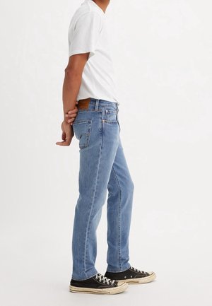 Man standing sideways wearing light blue jeans, white t-shirt, and black sneakers with white soles, hands clasped behind back.