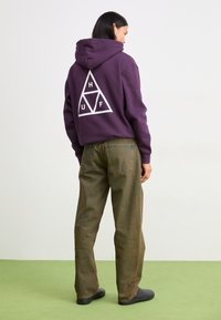 Purple hooded sweatshirt with a white triangular graphic on the back, paired with loose brown denim jeans and black slip-on shoes.