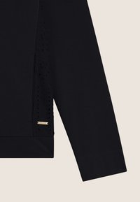 Top negro de manga larga con un panel lateral texturizado con recortes florales y un pequeño logo dorado. Tela suave, ajuste relajado.