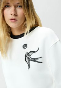 Felpa bianca con dettagli neri, caratterizzata da un disegno di rondine e un logo a forma di cuore sul petto. Texture liscia, vestibilità casual.