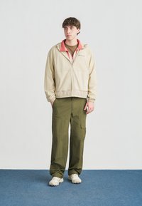 Junger Mann steht und trägt eine beige Reißverschlussjacke über einem korallenfarbenen Kapuzenpullover, olivgrüne Cargohosen und weiße Turnschuhe auf einem blauen Teppich.