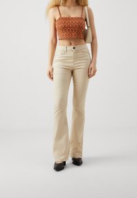 Mujer vestida con pantalones acampanados beige, tacones negros y un top corto de patrón naranja, llevando un bolso de hombro beige, de pie sobre un suelo blanco.
