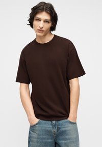 Νεαρό άτομο με μεσαίου μήκους σκούρα μαλλιά, φορώντας σκούρο καφέ κοντομάνικο μπλουζάκι και μπλε τζιν, με τα χέρια στις τσέπες, με ουδέτερη έκφραση.