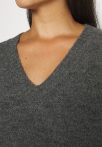 Gros plan sur une personne portant un pull en V doux de couleur gris foncé, montrant le col et la partie supérieure de la poitrine.