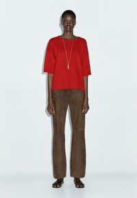 Maglione rosso lavorato a maglia con vestibilità comoda, abbinato a pantaloni a gamba larga effetto suede marroni. L'outfit è completato da una lunga collana con ciondolo dorato.