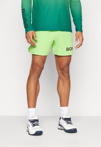 Lime žalios spalvos maudymosi šortai turi juodą "BORG" logotipą, pasižymi lengvu dizainu ir greitai džiūstančia medžiaga, derinami su baltais ir tamsiai mėlynais sportbačiais.
