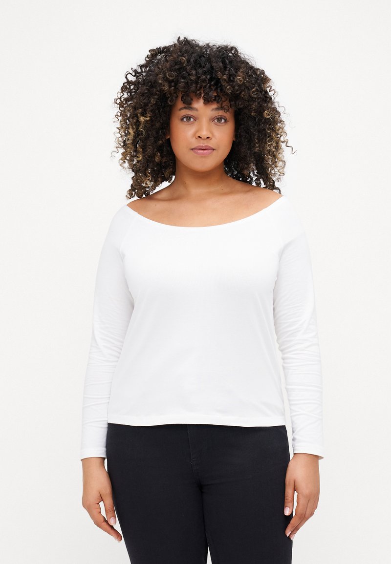Top blanco de manga larga con escote off-the-shoulder, hecho de material suave. Textura lisa, combinado con jeans oscuros.