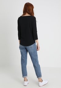 Schwarzes Langarmoberteil, kombiniert mit hellblauen Jeans mit umgekrempelten Bündchen und weißen Sneakers. Model von hinten mit glattem Haar gezeigt.