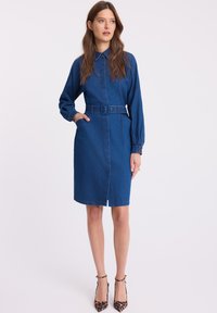 Sukienka koszulowa z denimu, niebieska, w talii z paskiem, z długimi rękawami, kołnierzykiem, z dwiema kieszeniami, do kolan, z guzikami na mankietach. Spodnie nie są pokazane.