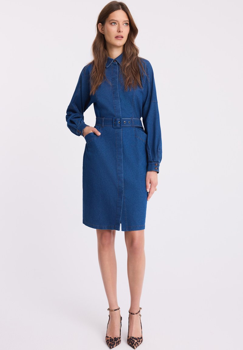 Sukienka koszulowa z denimu, niebieska, w talii z paskiem, z długimi rękawami, kołnierzykiem, z dwiema kieszeniami, do kolan, z guzikami na mankietach. Spodnie nie są pokazane.