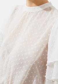 Blusa bianca trasparente con pois testurizzati, maniche arricciate e scollo alto. Tessuto leggero con un aspetto delicato e arioso.