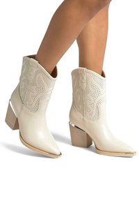 Białe skórzane kowbojki za kostkę z wycięciami, spiczastym czubkiem i ozdobnym stębnowaniem. Posiadają drewnianą podeszwę z metalicznym akcentem.