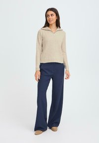 Beige strikkegenser med glidelås i kragen, kombinert med marineblå brede bukser. Stoffet ser mykt ut og har en vertikal ribbestruktur.