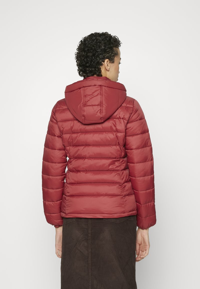 Persona con cabello rizado que lleva una chaqueta acolchada con capucha roja y una falda marrón, vista desde atrás sobre un fondo sencillo.