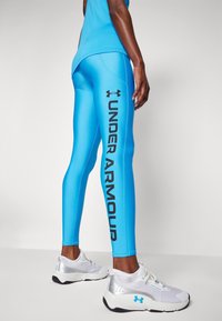 Osoba ubrana w jasnoniebieskie legginsy Under Armour z logo napisanym wzdłuż nogawki oraz białe sportowe buty z niebieskimi akcentami.