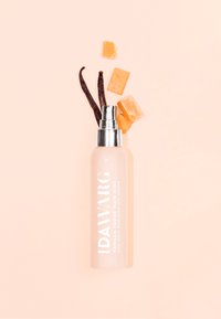 Przezroczysta butelka spray z metalową pompką, oznaczona etykietą "IDAWARG Vanilla Fudge Hair Mist", otoczona laskami wanilii i kawałkami karmelu na miękkim różowym tle.