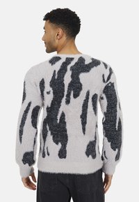 Pull en peluche blanc avec des motifs abstraits noirs, présentant un col rond et des manches longues. Texture douce, coupe décontractée et poignets profonds.