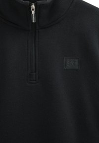 Schwarzer Pullover mit einem halbleichtem Kragen, strukturiertem Stoff und einem rechteckigen schwarzen Patch mit der Aufschrift "POLO CLUB", auf der Brust genäht.