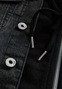 Giacca di jeans nera con accessori in metallo argentato, caratterizzata da bottoni automatici, cerniera e cordino nero con estremità brandizzate. Dettagli in tessuto testurizzato visibili.