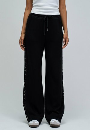 Persona con pantalones negros de pierna ancha con cintura ajustable con cordón y franjas laterales decorativas, camiseta sin mangas blanca y zapatillas blancas, de pie sin moverse.