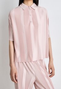 Ljusrosa plisserad polo med kort ärm och tre knappar, matchat med plisserade byxor för en koordinerad look.
