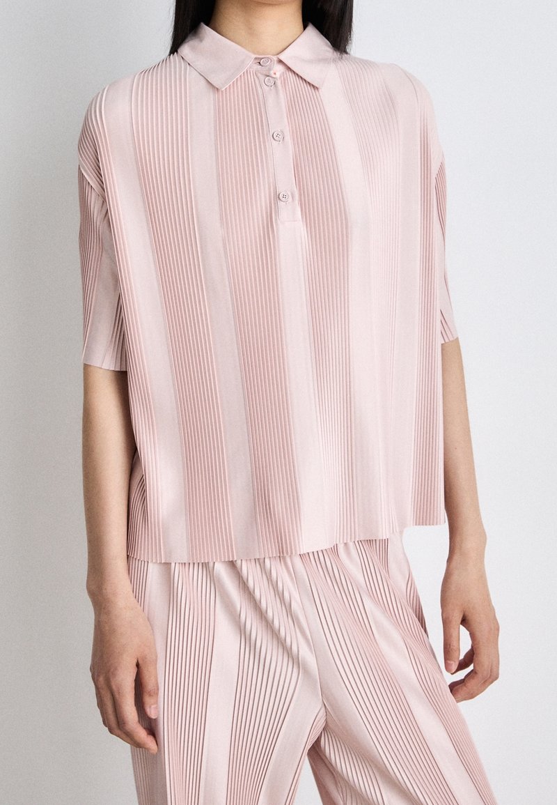 Ljusrosa plisserad polo med kort ärm och tre knappar, matchat med plisserade byxor för en koordinerad look.